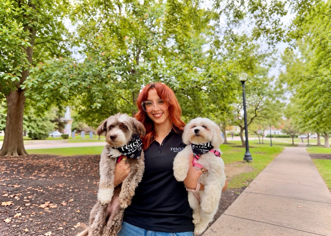 A smiling Fetch! Pet Care sitter holding two small dogs in Fetch! bandanas in a tree-lined park
