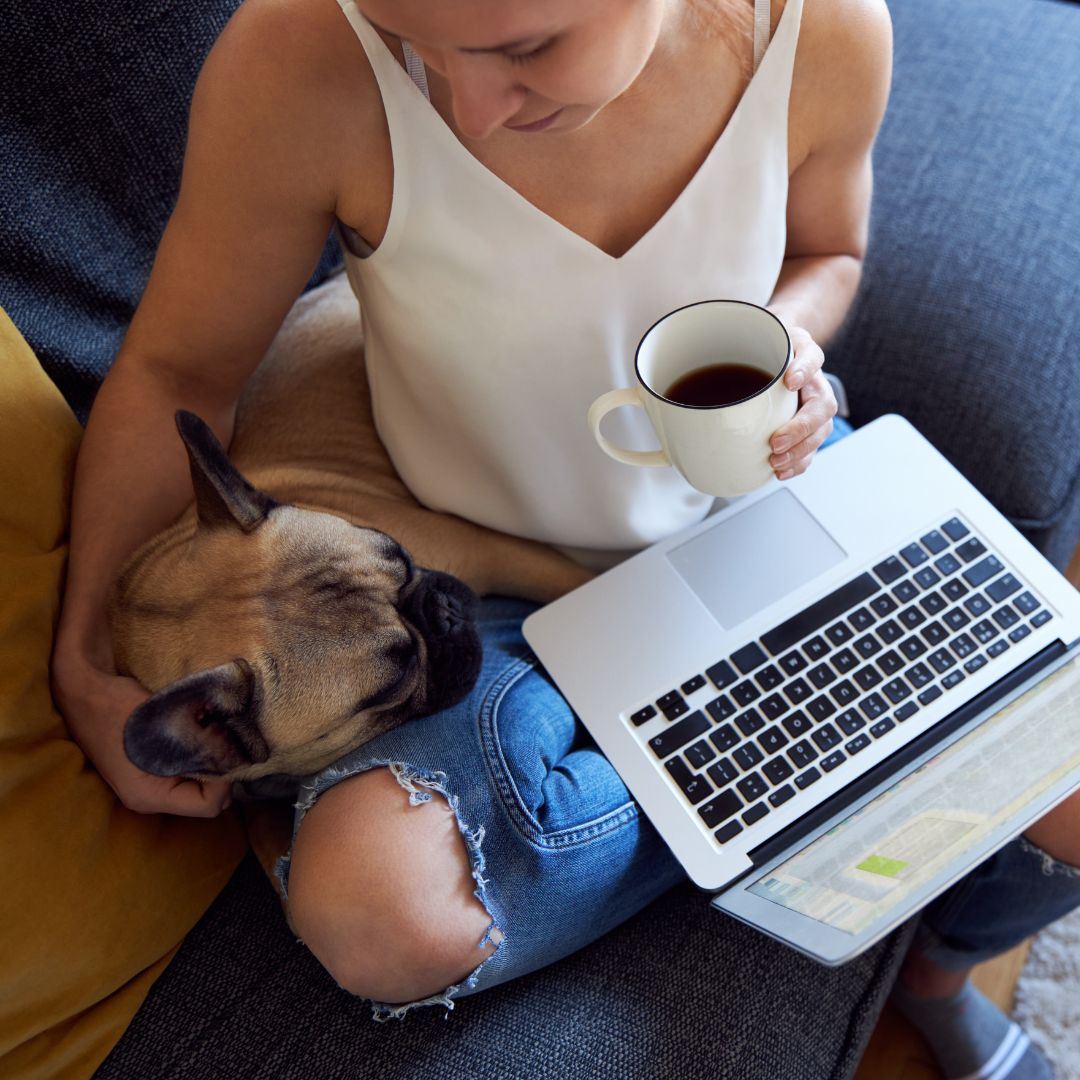 Person using a laptop and holding a cup of coffee with their dog sleeping by their side